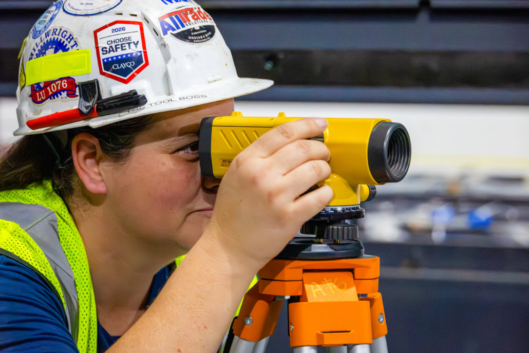 3T5A3553-2 Female Union Millwright wearing hard hat peering through sight to assess precision level on equipment.